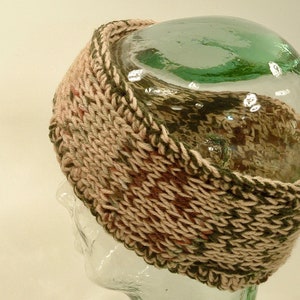 May include: A knitted headband in a brown, green, and beige color pattern. The headband is displayed on a glass mannequin head.