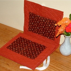 May include: A pair of orange and brown quilted fabric placemats with a floral pattern. The placemats are folded in half and have a white string attached to them.