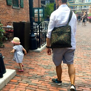 May include: A man wearing a white shirt and blue shorts walks on a brick pathway with a floral patterned shoulder bag. A young girl wearing a pink hat and blue dress walks in the opposite direction.