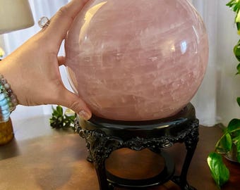May include: A large, polished rose quartz sphere, resting on a black, ornate stand. The sphere is a soft pink color with natural veining. The stand has a detailed, scrolled design.