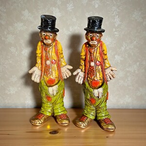 May include: Two vintage clown figurines. Each clown wears a black top hat, yellow jacket, orange vest, and green pants. The clowns have a sad expression and open hands. The shoes are gold. The figurines are on a wooden surface.