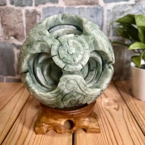 May include: A carved jade sphere with intricate floral and geometric designs, resting on a wooden stand. The sphere is a mix of green hues, and the stand is a light brown color. The background features a brick wall and a plant.