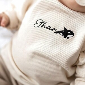 May include: Cream-colored baby long-sleeved shirt with the name "Ethan" in black script and a black and white orca whale design. The shirt is made of ribbed fabric.