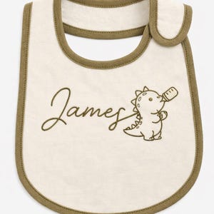 Puede incluir: Babero de bebé color crema con ribete marrón y el nombre "James" bordado en cursiva. Un dinosaurio de dibujos animados bebiendo de un biberón también está bordado en el babero. El texto "Font Included" está en la parte inferior.