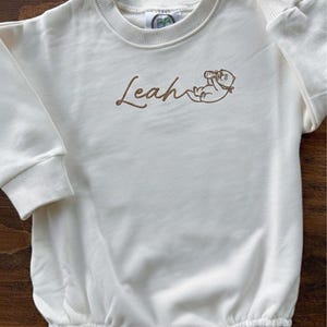 May include: Cream-colored baby romper with long sleeves and a round neckline. The name "Leah" is embroidered in brown script, along with a small illustration of a cat. The romper has snap closures at the bottom and is made of a soft fabric.