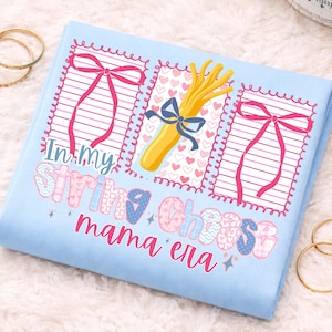 以下が含まれることがあります： 「In my string cheese mama era」のテキストが特徴の、グラフィックデザインが施された水色のTシャツ。ピンクのリボン、ストリングチーズのイラスト、ハート模様がデザインに含まれています。