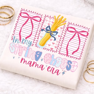 以下が含まれることがあります： 「In my string cheese mama era」の文字が入ったクリーム色のスウェットシャツ。ピンクのリボン、青いリボンで結ばれた黄色のストリングチーズの束、ハートのデザイン。背景には金のブレスレットがあります。