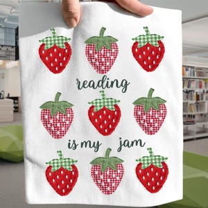 Puede incluir: Tela blanca con fresas bordadas y el texto "reading is my jam". Las fresas son rojas con tapas de cuadros verdes, algunas con un patrón de cuadros rojos y blancos. El texto está en una fuente cursiva.