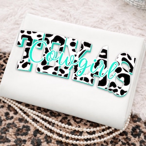 Puede incluir: Un objeto rectangular blanco con las palabras "Texas Cowgirl" en un diseño de estampado de vaca en blanco y negro con un contorno turquesa. El objeto está sobre una superficie con estampado de leopardo con perlas.