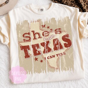 Puede incluir: Camiseta color crema con un gráfico desgastado con el texto "She's from TEXAS I CAN TELL" en rojo y un contorno del mapa de Texas. La camiseta tiene mangas cortas y cuello redondo.