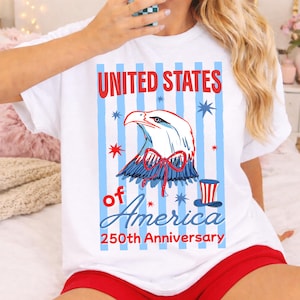 Puede incluir: Camiseta blanca con un diseño patriótico. El gráfico presenta un águila calva con detalles en rojo, blanco y azul, el texto "UNITED STATES of AMERICA 250th Anniversary" y rayas azules y blancas.