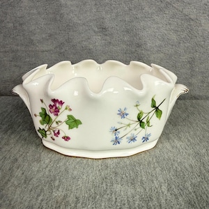 May include: An oval ceramic bowl with a ruffled, scalloped rim and two side handles. The white surface features painted botanical illustrations of purple-pink flowers with green leaves on one side and small blue flowers with green stems on the other.