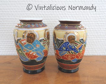 May include: Two small, decorative ceramic vases with a gold trim and intricate hand-painted designs of people in traditional Japanese clothing. The vases are on a wooden surface.