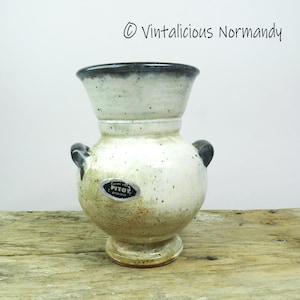 Pode incluir: Um vaso de cerâmica salpicada com uma parte superior alargada e duas pequenas alças. O vaso tem um corpo creme com uma borda e alças azul escuro. Uma pequena etiqueta com o texto "PITOT" está afixada ao vaso.