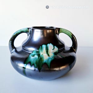 May include: A dark, metallic vase with two handles and a wide opening. The vase features a glossy finish with green and white floral accents. The design is reminiscent of Art Deco or Art Nouveau styles.