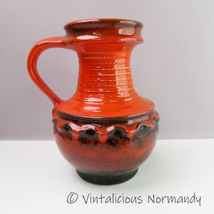 May include: A vintage ceramic pitcher with a handle, featuring a vibrant orange glaze with dark brown accents. The pitcher has a bulbous base and a ridged neck, with a decorative wavy pattern near the base. The base is black.