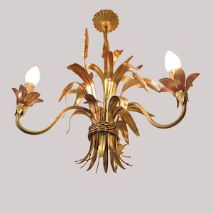 May include: Gold-toned metal chandelier with a wheat sheaf design. The fixture has three curved arms, each holding a light bulb. The central design features wheat stalks and leaves, with a woven band at the base.