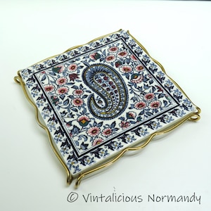 May include: Square ceramic tile with a floral and paisley design in shades of blue, pink, and white. The tile is set in a gold-toned metal frame with a wavy edge. The tile features a central paisley motif.