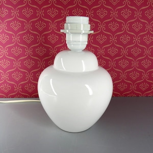 Op de afbeelding: Een witte keramische tafellampvoet met een gladde, afgeronde, hartvormige silhouet. De lamp is voorzien van een witte kunststof fitting met schroefdraad aan de bovenzijde voor een lamp en bevat een wit elektrisch snoer aan de onderkant.