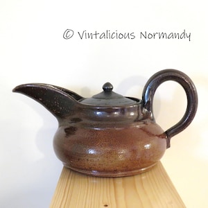 Può includere: Una teiera in ceramica marrone con coperchio scuro e beccuccio curvo. La teiera ha un corpo arrotondato e un grande manico ad arco. Il testo "© Vintalicious Normandy" è visibile nell'angolo in alto a destra.