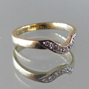 Op de afbeelding: Een goudkleurige ring met een gebogen ontwerp, bezet met kleine, heldere edelstenen. De band van de ring heeft een gepolijste afwerking en de edelstenen zijn langs het gebogen gedeelte gerangschikt. De ring wordt weerspiegeld in een glanzend oppervlak.
