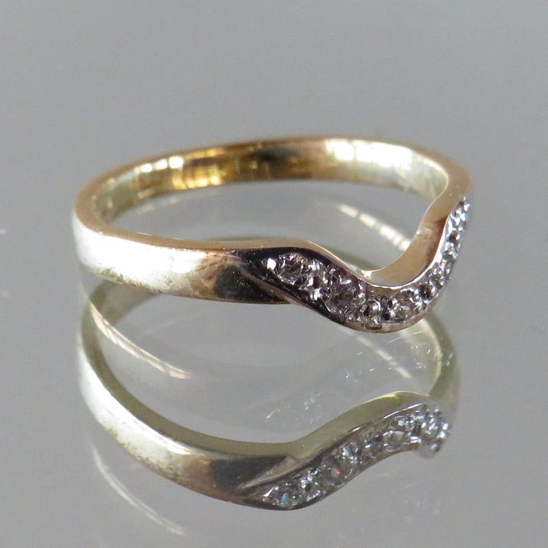 Op de afbeelding: Een goudkleurige ring met een gebogen ontwerp, bezet met kleine, heldere edelstenen. De band van de ring heeft een gepolijste afwerking en de edelstenen zijn langs het gebogen gedeelte gerangschikt. De ring wordt weerspiegeld in een glanzend oppervlak.