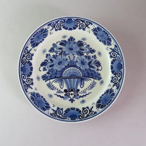 Può includere: Un piatto rotondo in ceramica con base bianca e dettagliati motivi floreali dipinti a mano in blu. Il centro mostra un grande bouquet di fiori in un'urna decorativa, mentre il bordo è ornato da volute e motivi floreali blu ripetuti.