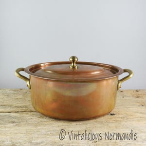 May include: A copper pot with brass handles and a lid. The pot has a tarnished finish and is sitting on a wooden surface.