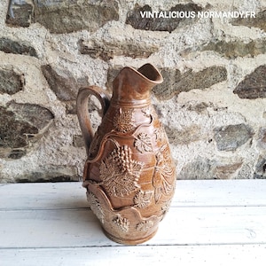 Op de afbeelding: Een bruine keramische kan met een handvat en een tuit. De kan is versierd met reliëf druivenranken en bladeren. De tekst "VINTALICIOUS NORMANDY.FR" is rechtsboven zichtbaar. De kan staat op een wit oppervlak.