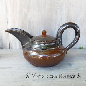 Può includere: Una teiera in ceramica marrone con coperchio e manico curvo. La teiera ha un beccuccio e una finitura lucida. Il testo "© Vintalicious Normandy" è in basso nell'immagine.