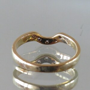 Op de afbeelding: Een goudkleurige ring met een gebogen ontwerp, met verschillende kleine, heldere edelstenen langs de bovenste curve. De ring wordt weerspiegeld in een glanzend oppervlak, waardoor een spiegeleffect ontstaat. De ringband is glad en gepolijst.