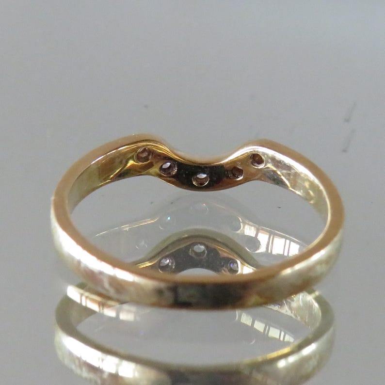 Op de afbeelding: Een goudkleurige ring met een gebogen ontwerp, met verschillende kleine, heldere edelstenen langs de bovenste curve. De ring wordt weerspiegeld in een glanzend oppervlak, waardoor een spiegeleffect ontstaat. De ringband is glad en gepolijst.