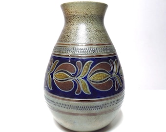 May include: A ceramic vase with a speckled tan top and a blue band featuring a floral design in brown and yellow. The vase has a rounded body and a narrow neck. The base is a light grey colour.