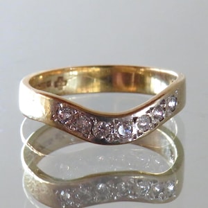 May include: A curved gold ring with a row of small, clear gemstones set along the top. The ring is reflected in the shiny surface below. The ring's band is a warm gold color, and the gemstones sparkle.