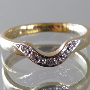 Op de afbeelding: Een goudkleurige ring met een gebogen ontwerp, bezet met kleine, heldere edelstenen. De band van de ring heeft een gladde, gepolijste afwerking en de edelstenen zijn langs de gebogen bovenkant gerangschikt. De ring wordt op een reflecterend oppervlak weergegeven.