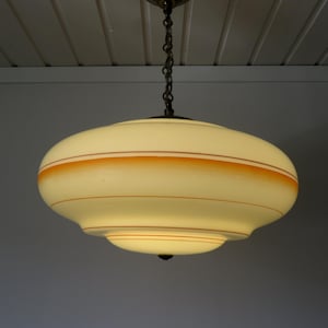Può includere: Una lampada a sospensione con un ampio paralume in vetro a forma di piattino di colore bianco crema. Il paralume è decorato con strisce orizzontali arancioni ed è appeso al soffitto tramite una catena in metallo scuro e un'asta centrale.