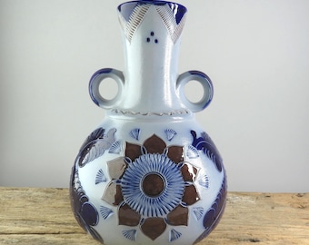 May include: A ceramic vase with a light blue base, decorated with dark blue and brown floral patterns. It has two loop handles and a detailed neck with a geometric design. The vase is a decorative item.