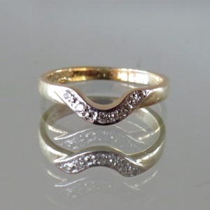 Op de afbeelding: Een goudkleurige ring met een gebogen ontwerp, bezet met kleine, heldere edelstenen. De band van de ring heeft een gladde, gepolijste afwerking en de edelstenen zijn geclusterd langs de gebogen bovenkant. De ring wordt weerspiegeld in het oppervlak eronder.