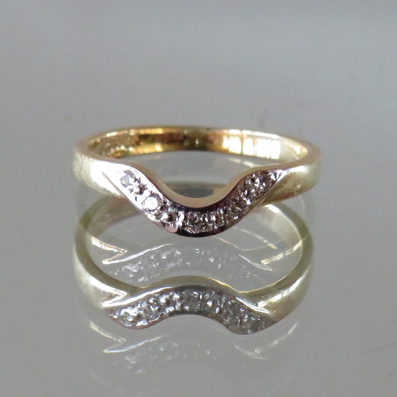 Op de afbeelding: Een goudkleurige ring met een gebogen ontwerp, bezet met kleine, heldere edelstenen. De band van de ring heeft een gladde, gepolijste afwerking en de edelstenen zijn geclusterd langs de gebogen bovenkant. De ring wordt weerspiegeld in het oppervlak eronder.