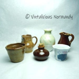 May include: A collection of six small ceramic pottery pieces, including a brown and tan pitcher, a brown and tan bowl, a brown bottle with a cork stopper, a green and white vase, a brown pitcher with a handle, and a white cup with a blue and pink floral design.