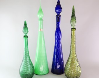 May include: Four tall glass decanters with stoppers. The first is emerald green with a hobnail texture. The second is a smooth, light mint green. The third is deep cobalt blue with a grid pattern. The fourth is olive green with a hobnail texture.