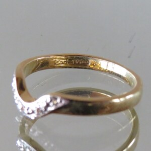 Op de afbeelding: Een goudkleurige ring met een gebogen ontwerp. E&eacute;n kant van de ring is bezet met kleine, heldere edelstenen. De ring heeft een gladde, gepolijste afwerking en een keurmerk. De ring wordt gepresenteerd op een reflecterend oppervlak.