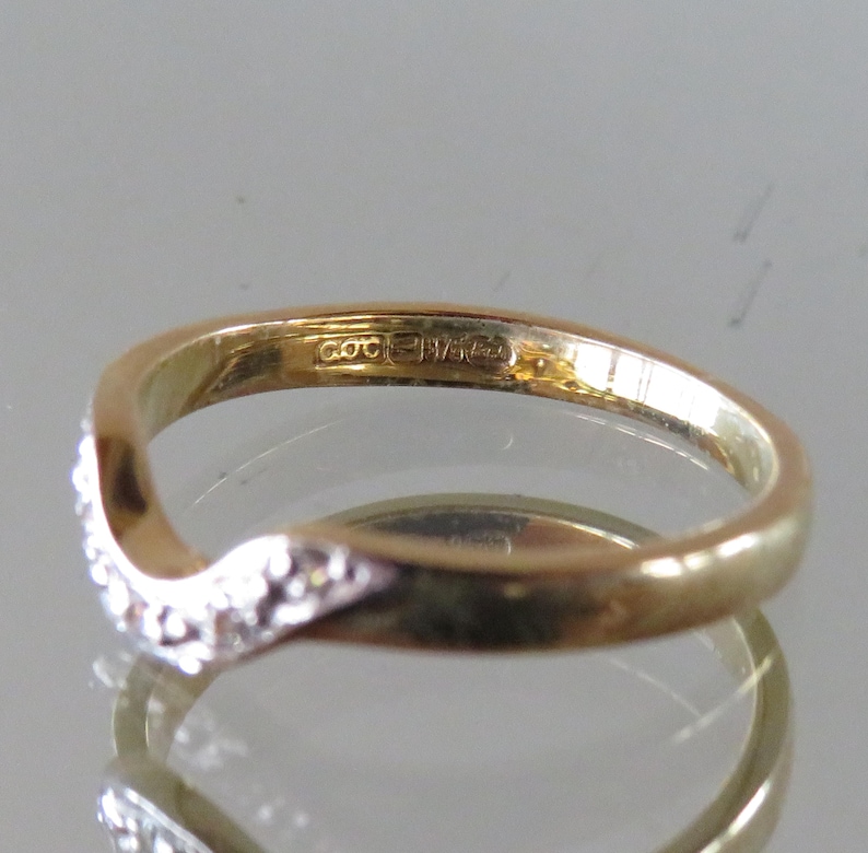 Op de afbeelding: Een goudkleurige ring met een gebogen ontwerp. E&eacute;n kant van de ring is bezet met kleine, heldere edelstenen. De ring heeft een gladde, gepolijste afwerking en een keurmerk. De ring wordt gepresenteerd op een reflecterend oppervlak.