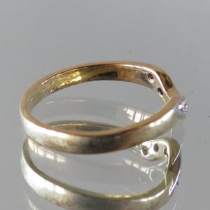 Op de afbeelding: Een goudkleurige ring met een gebogen ontwerp, met kleine, heldere edelstenen. De ring heeft een gepolijste afwerking en wordt gepresenteerd op een reflecterend oppervlak, wat de vorm en glans benadrukt. Het ontwerp van de ring is elegant en eenvoudig.