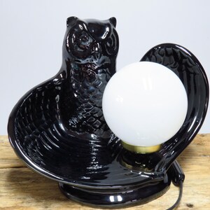 May include: A black ceramic owl-shaped lamp with a white globe light. The lamp has a glossy finish and a gold-colored base for the light. The owl's wings form a cradle for the light.