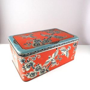 May include: Rectangular red tin box with a blue border and floral and bird designs. The box features a textured surface and a lid. The artwork includes white and blue flowers and birds.