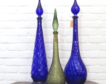 May include: Three decorative glass bottles. Two are cobalt blue with spiral and diamond patterns, and the third is olive green with a textured surface. Each bottle has a unique stopper design. They are displayed on a wooden table.
