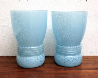 May include: Two light blue vintage drinking glasses with a speckled design. The glasses have a bulbous base and a cylindrical top. The glasses are made of glass and are sitting on a wooden surface.