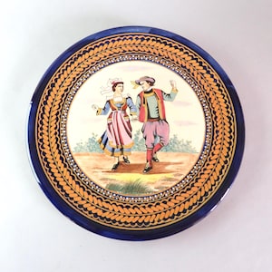 May include: A round ceramic decorative plate featuring a central illustration of two figures in traditional attire dancing on a light brown ground. The plate has a wide dark blue border with an intricate orange braided pattern and white dots.