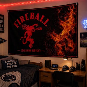 K&ouml;nnte beinhalten: Ein schwarzes Fireball Cinnamon Whisky Banner mit roter Teufelsgrafik und Flammen. Der Raum umfasst einen Schreibtisch mit Digitaluhr, Penn State Kappen und einem Neon-Schild "Let's Go!". Der Raum ist mit roten Lichtern beleuchtet.